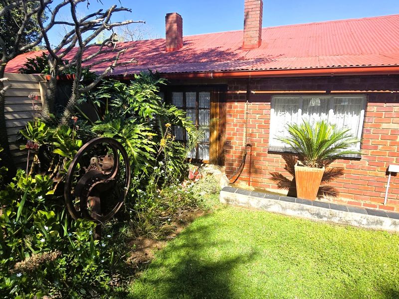 Rear view of a brick house featuring a corrugated metal roof, brick chimneys, and a small garden area with established landscaping.