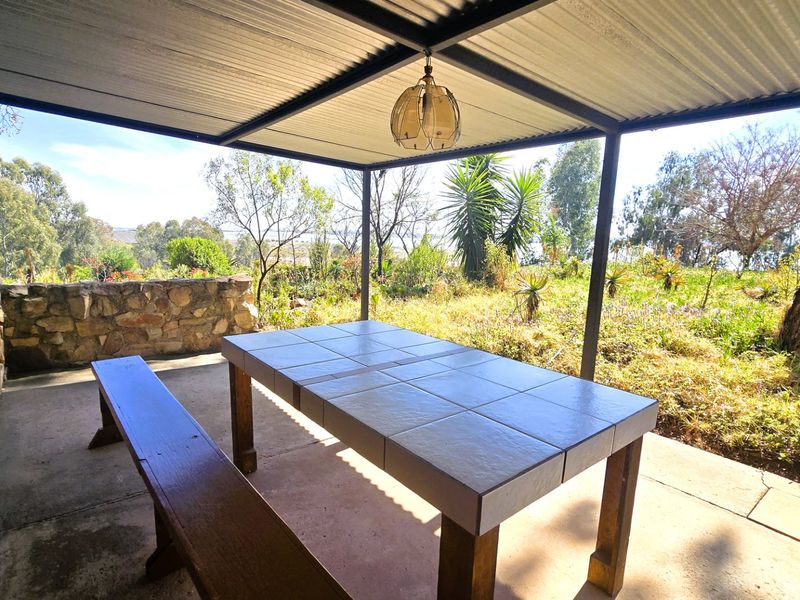 Covered outdoor patio featuring a corrugated metal roof, stone wall accents, and concrete flooring, overlooking a natural garden setting.