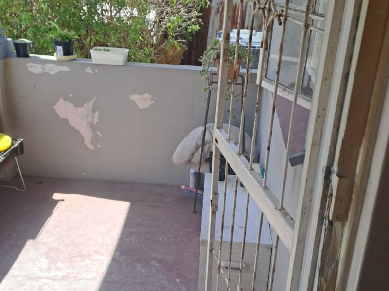 A small outdoor balcony area featuring a metal security gate and concrete flooring, requiring some maintenance to the wall finishes and gate.