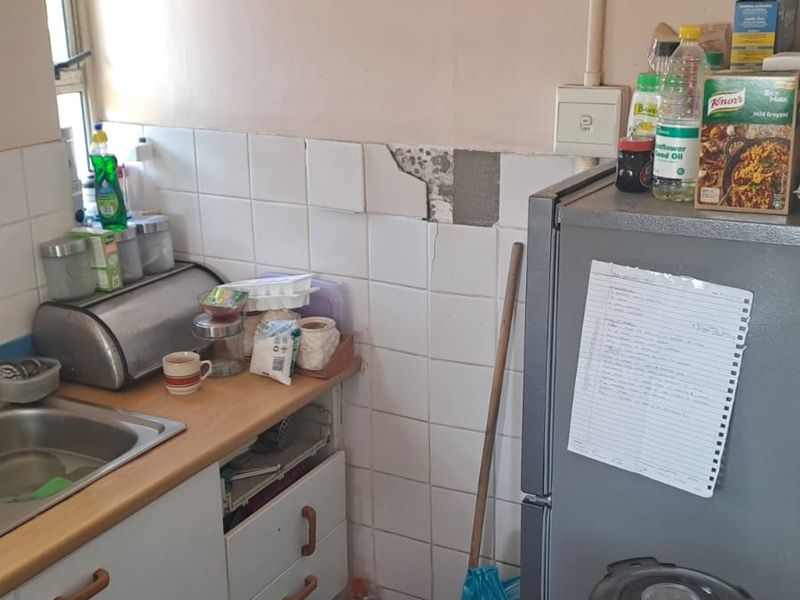 A compact kitchen area featuring a stainless steel sink, laminate countertops, and tiled splashbacks, currently showing signs of wear and missing wall tiles.