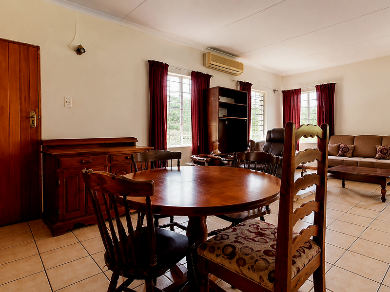 Spacious open-plan living and dining area featuring ceramic tile flooring, a wall-mounted air conditioning unit, and large windows with security bars.