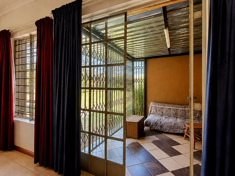 A covered patio area featuring tiled flooring, a corrugated metal roof, and a security gate, providing a transition space to the outdoor garden.