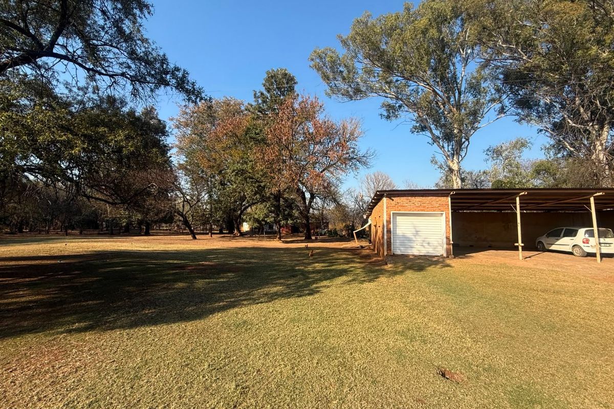 A spacious backyard featuring a large lawn, mature trees, a brick garage, and an attached carport structure.