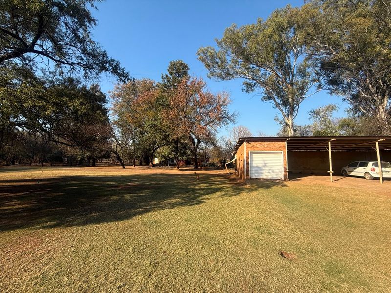 A spacious backyard featuring a large lawn, mature trees, a brick garage, and an attached carport structure.