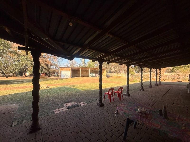 A spacious covered patio featuring brick paving and decorative timber columns, overlooking a large garden area with an additional detached garage structure.