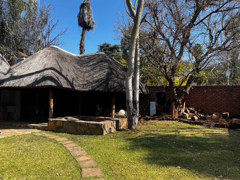 Outdoor entertainment area featuring a traditional thatch roof lapa with a stone-clad built-in braai, set within a private garden with brick boundary walls.
