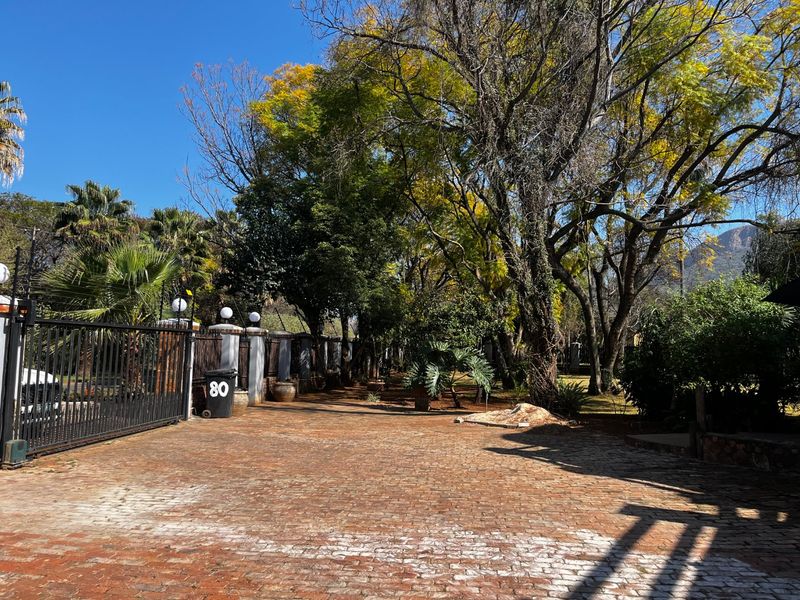 A spacious brick-paved driveway leading to the property entrance, featuring mature trees, perimeter walling, and a secure metal gate.