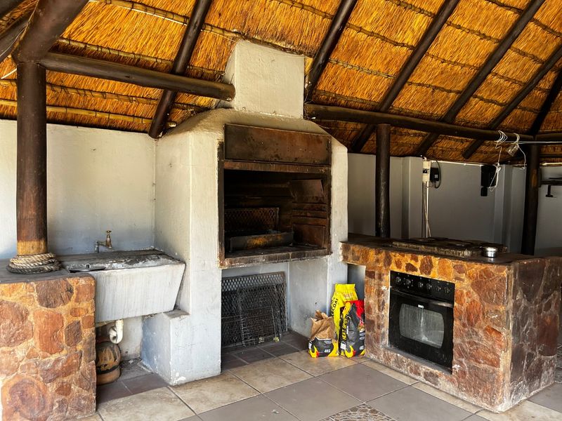 Covered outdoor entertainment area featuring a traditional thatched roof, built-in braai, stone-clad prep counter with integrated oven, and a utility sink.