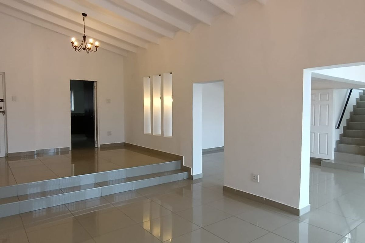 Spacious, open-plan living area featuring polished tiled floors, a raised platform, exposed beam ceilings, and a staircase leading to the upper level.