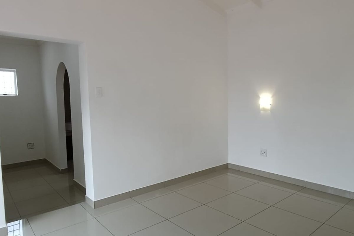 A bright, empty bedroom featuring neutral tiled flooring, wall-mounted lighting, and an integrated air conditioning unit, leading through an archway to an adjacent space.