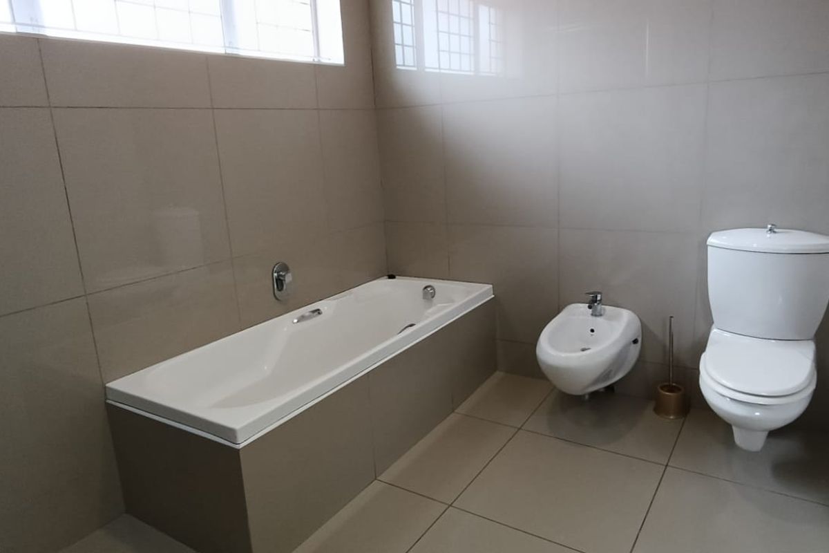 A clean, modern bathroom featuring a built-in bathtub, bidet, and toilet with neutral floor-to-ceiling wall tiles and a high-level window for natural light.