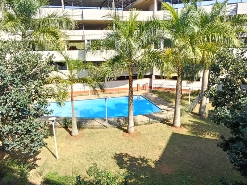 A communal outdoor area featuring a swimming pool, landscaped garden with palm trees, and paved walkways within the apartment complex.