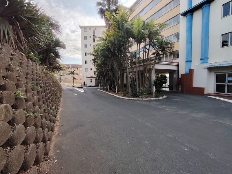 Exterior view of the apartment complex entrance featuring a paved driveway, covered drop-off area, and landscaped retaining wall.
