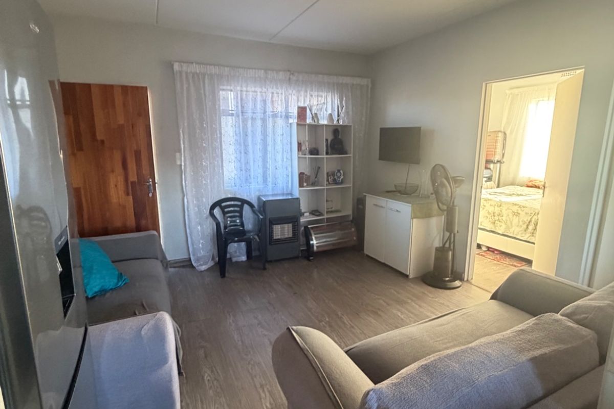 A living room area featuring laminate flooring and a wooden door, providing access to an adjacent bedroom.