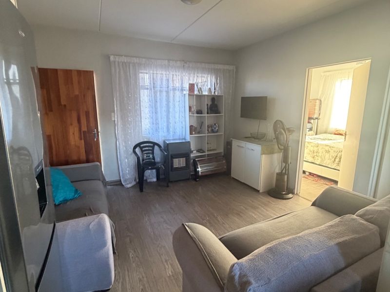A living room area featuring laminate flooring and a wooden door, providing access to an adjacent bedroom.