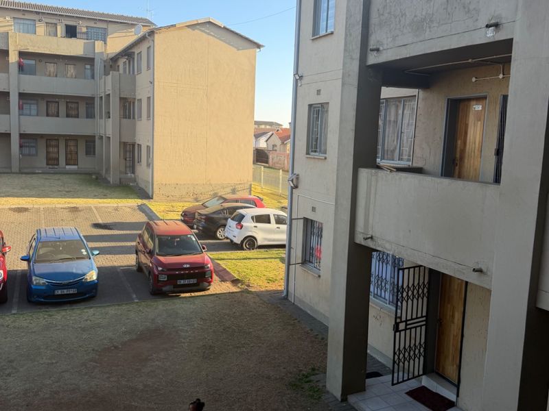 View of the apartment complex exterior, featuring a communal courtyard, parking area, and building facade with security gates and tiled entrance landings.