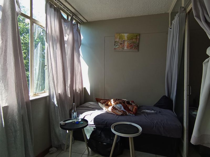 A bedroom featuring large windows that provide natural light, tiled flooring, and a textured ceiling. The space requires maintenance, including window repairs.