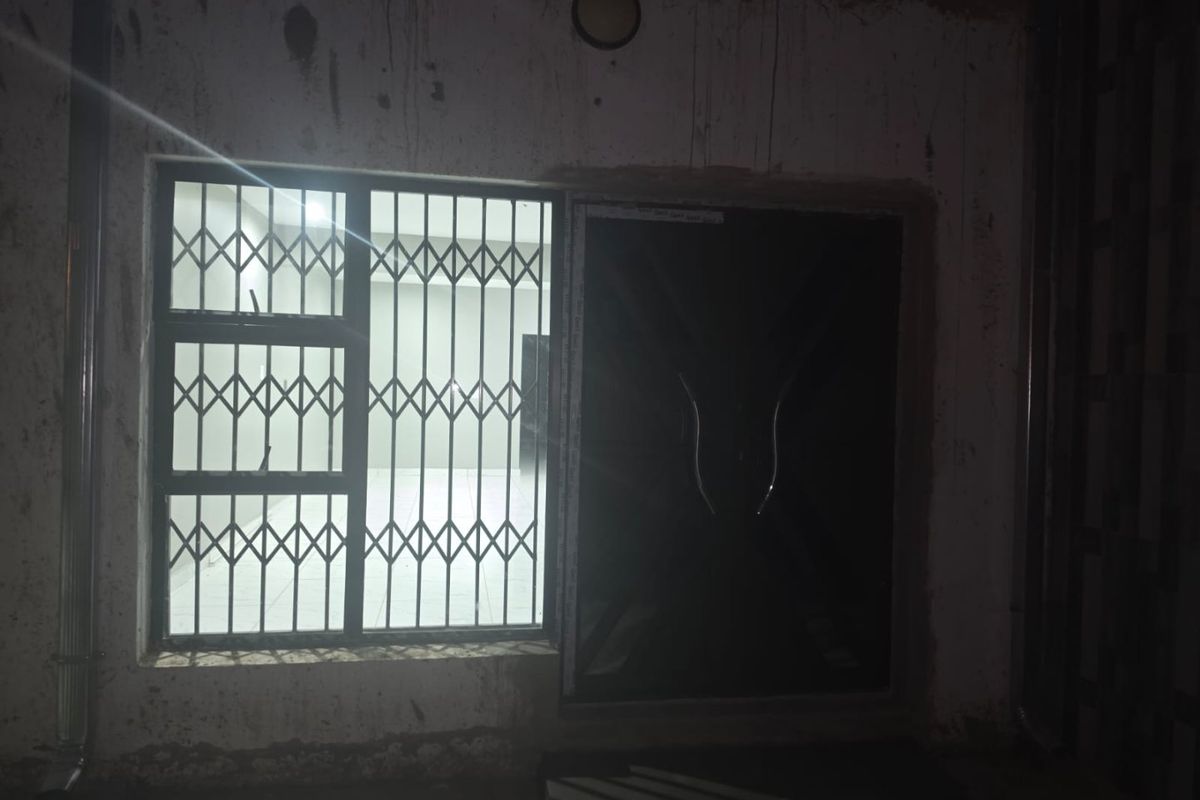 Exterior view of a rear building wall featuring a security gate and window with security bars, showing signs of unfinished plaster and wear.