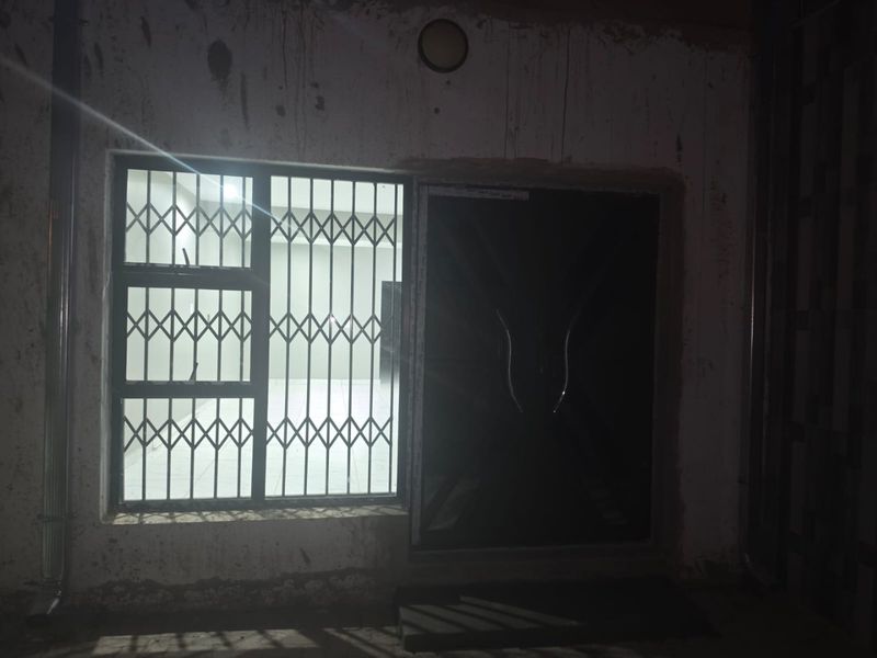 Exterior view of a rear building wall featuring a security gate and window with security bars, showing signs of unfinished plaster and wear.