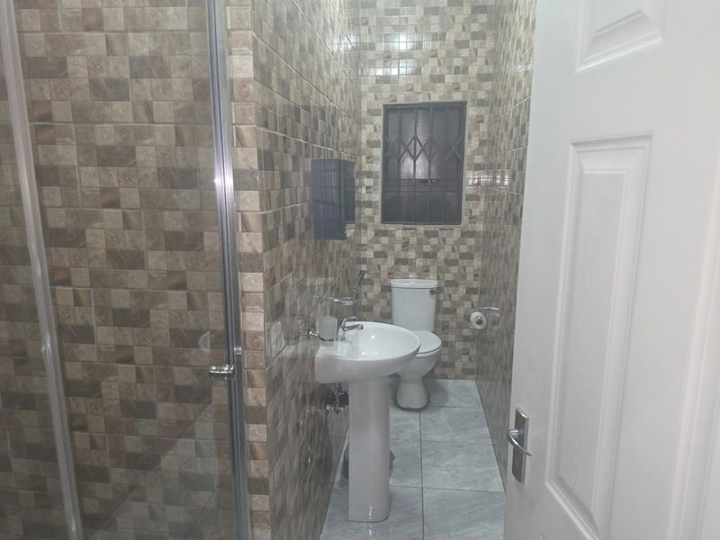 A compact bathroom featuring a pedestal basin, toilet, and a glass-enclosed shower, finished with full-height decorative wall tiling and tiled flooring.