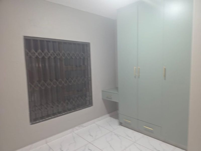 A clean, empty bedroom featuring modern light-colored built-in wardrobes, a matching vanity unit, and white marble-look tiled flooring with a security gate on the window.