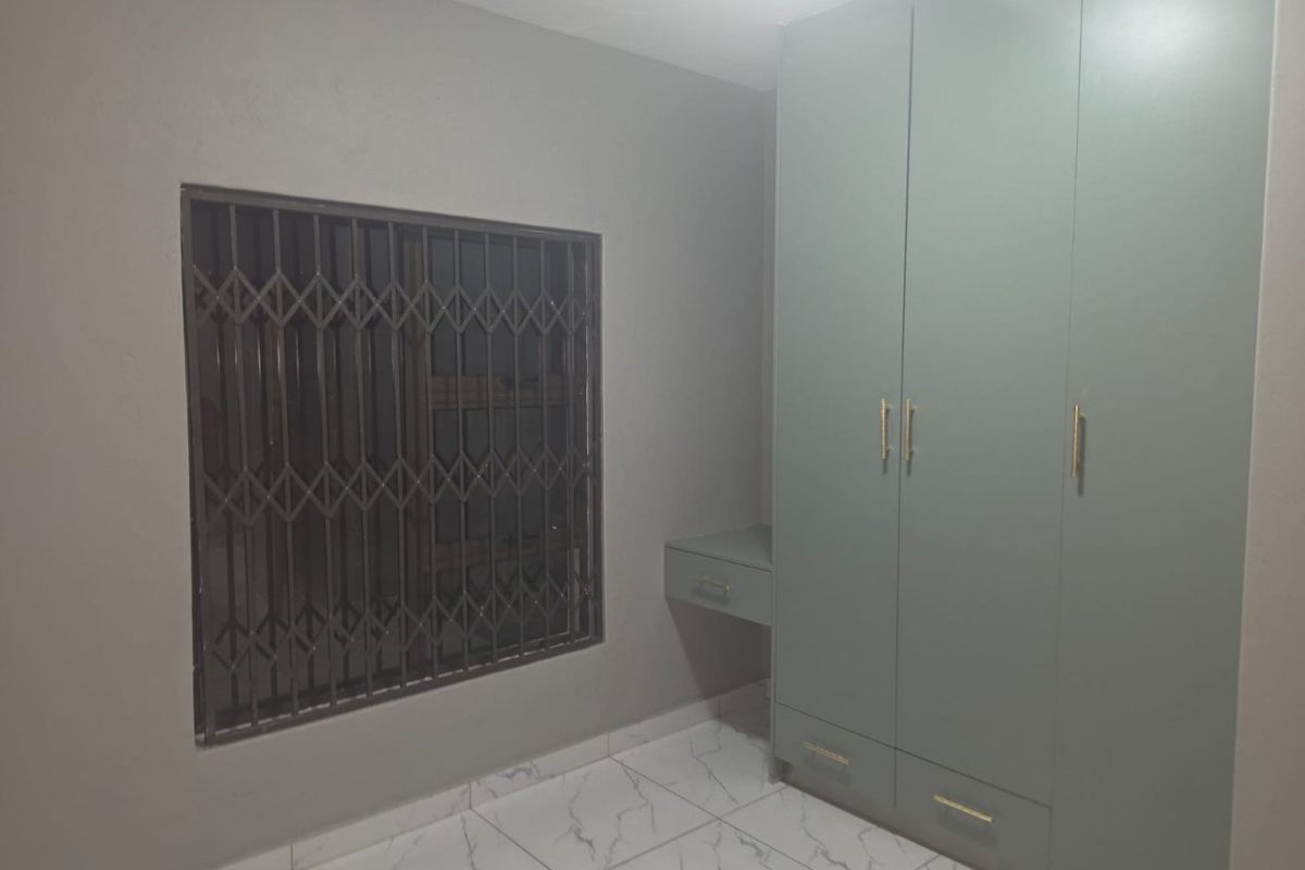 A clean bedroom featuring modern light green built-in wardrobes with a matching vanity, marble-look tiled flooring, and a window with a security gate.