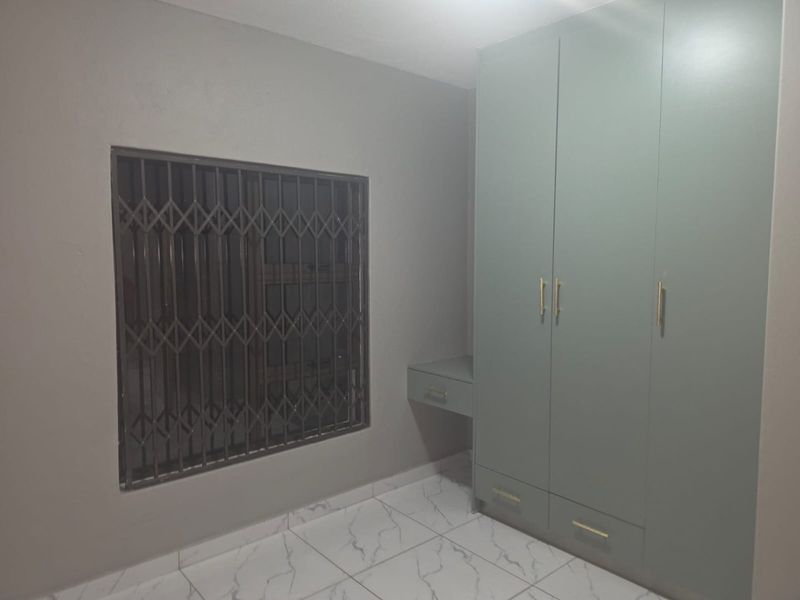 A clean bedroom featuring modern light green built-in wardrobes with a matching vanity, marble-look tiled flooring, and a window with a security gate.