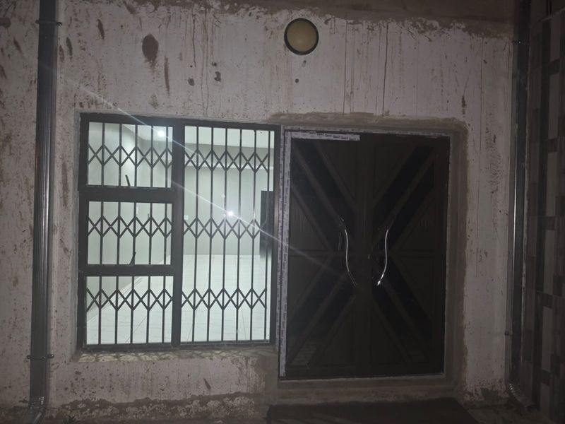 Exterior view of an apartment entrance featuring a dark security door and window with security bars, showing unfinished wall plaster and exterior staining.
