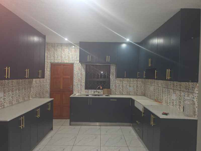 A modern kitchen featuring dark cabinetry, stone countertops, a double basin sink, and decorative tiled backsplash with neutral floor tiles.