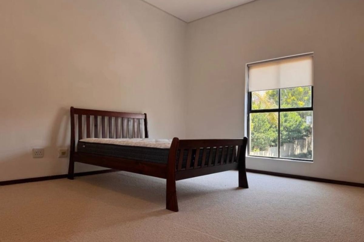 A bright and neutral bedroom featuring carpeted flooring, a large window with a fitted roller blind, and clean wall finishes.