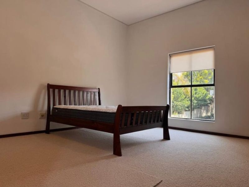 A bright and neutral bedroom featuring carpeted flooring, a large window with a fitted roller blind, and clean wall finishes.