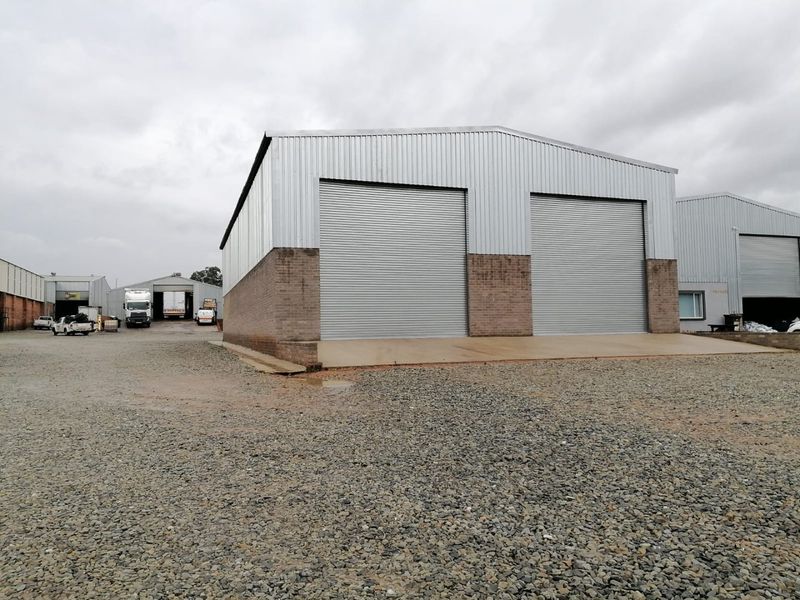 Industrial warehouse facility featuring dual metal roller shutter doors, corrugated metal cladding, and a spacious gravel yard for vehicle access.