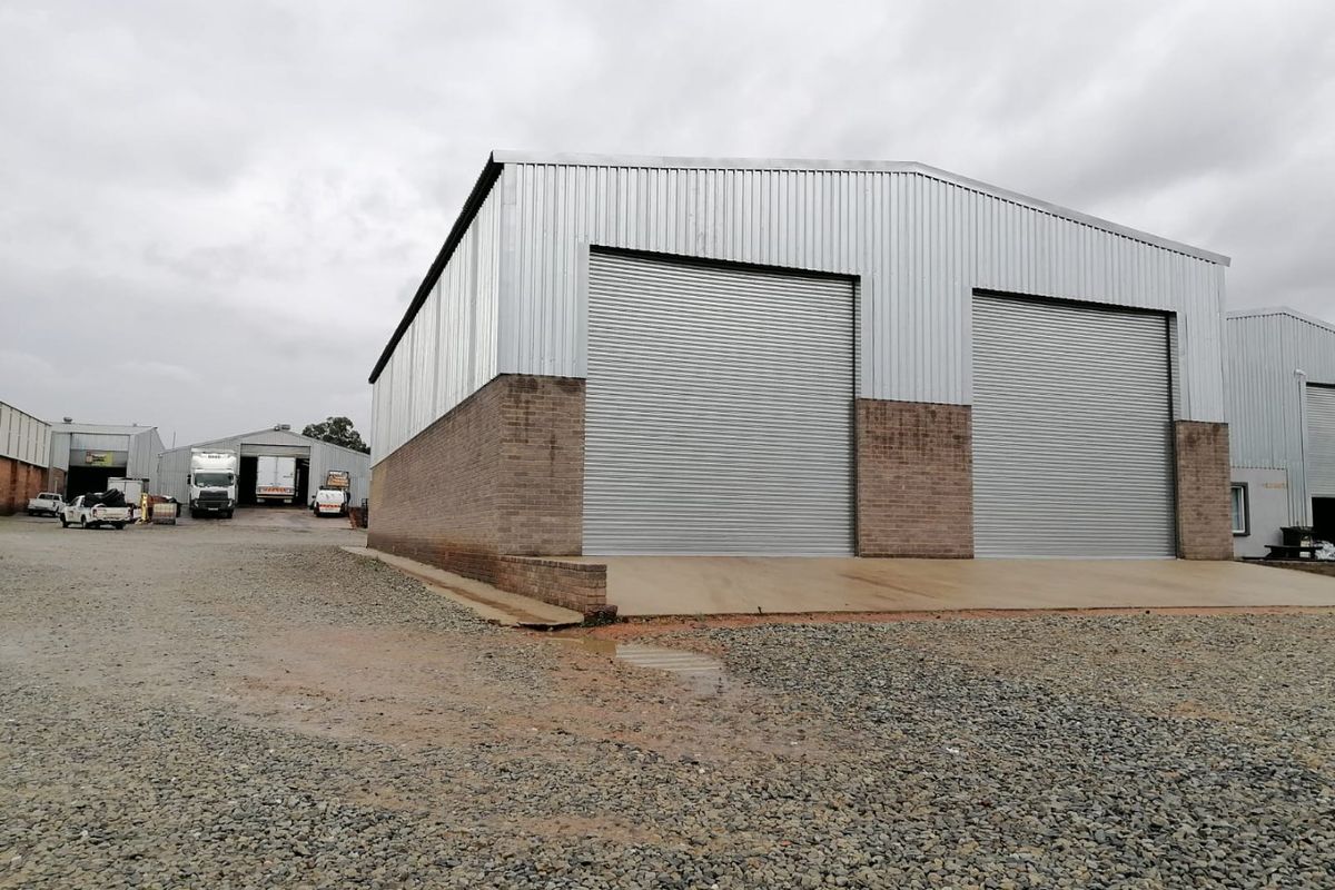 Industrial warehouse building featuring corrugated metal cladding, brick masonry base, and two large roller shutter doors with a concrete apron and gravel yard.
