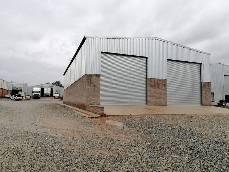 Industrial warehouse building featuring corrugated metal cladding, brick masonry base, and two large roller shutter doors with a concrete apron and gravel yard.
