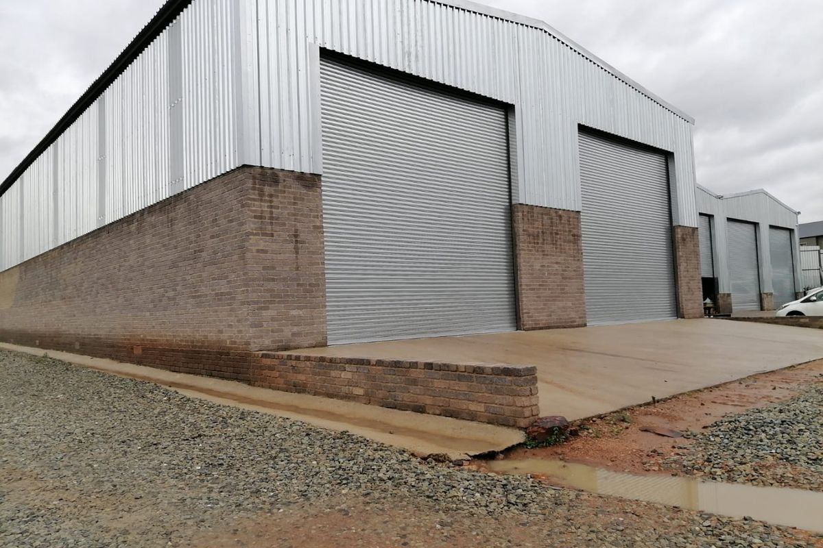 Industrial warehouse exterior featuring corrugated metal cladding, brick masonry base, and multiple large roller shutter doors with a concrete apron.