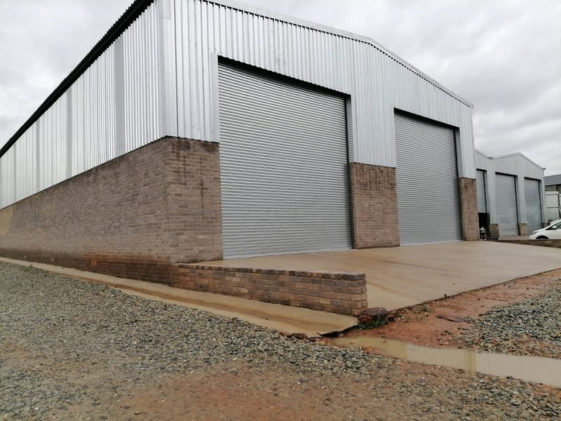 Industrial warehouse exterior featuring corrugated metal cladding, brick masonry base, and multiple large roller shutter doors with a concrete apron.