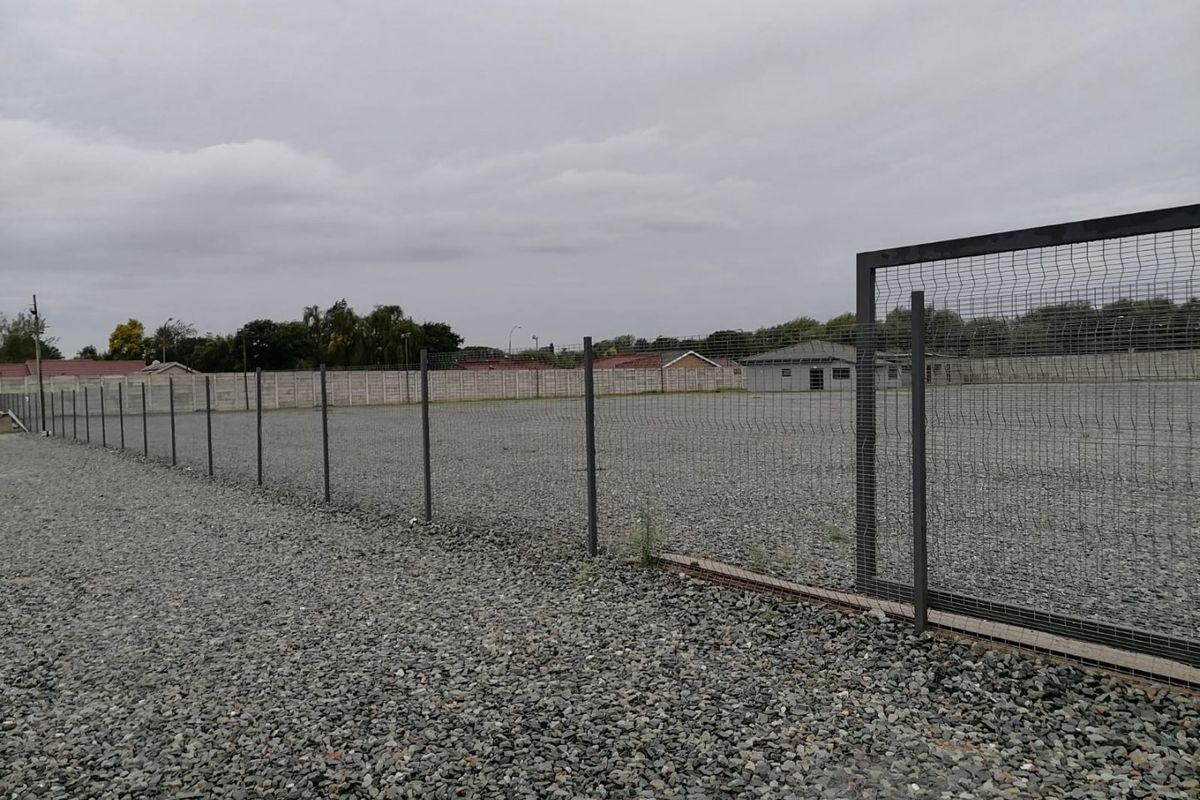 A spacious industrial yard featuring a gravel surface, enclosed by secure mesh perimeter fencing and a precast concrete boundary wall.