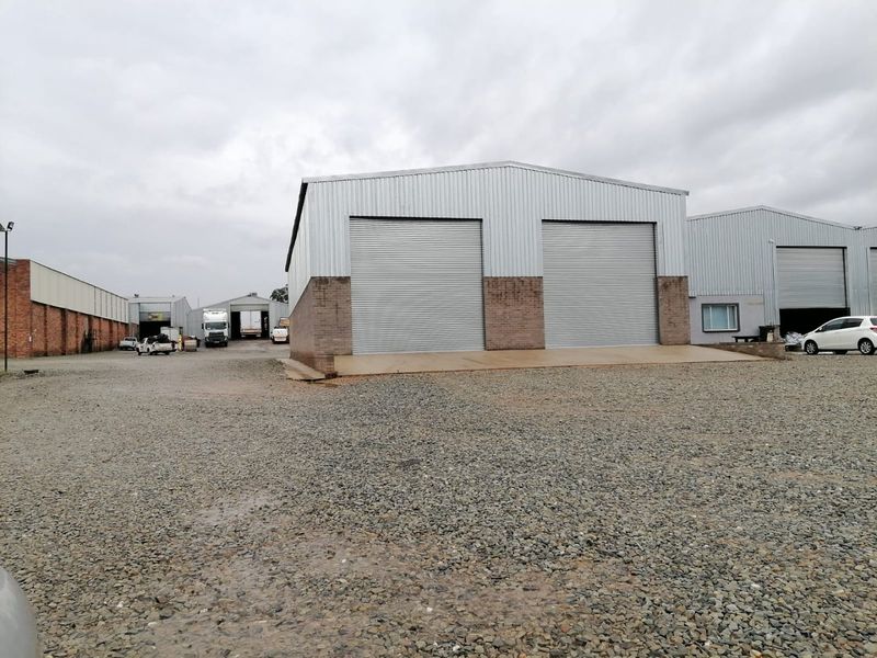 Industrial warehouse facility featuring dual roller shutter doors, brick and metal cladding construction, and a spacious gravel yard for vehicle access.