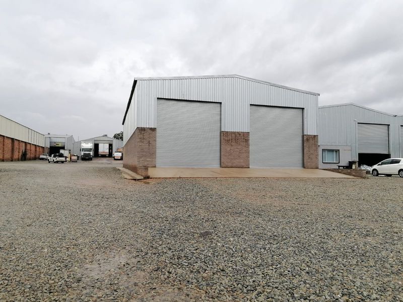 Industrial warehouse facility featuring dual metal roller shutter doors, corrugated metal cladding, and a spacious gravel yard for vehicle access.
