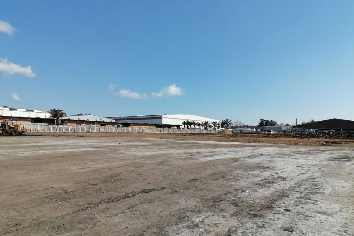 A wide-angle view of a cleared, level residential vacant land plot with perimeter fencing and surrounding industrial buildings in the background.