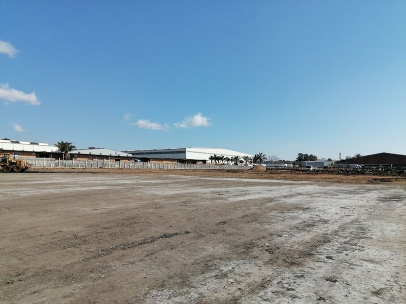 A wide-angle view of a cleared, level residential vacant land plot with perimeter fencing and surrounding industrial buildings in the background.