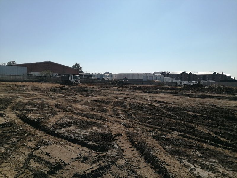 A wide-angle view of a cleared, vacant residential plot showing earthworks and surrounding industrial and residential structures.