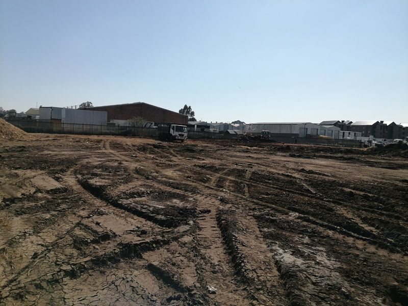 A vacant residential plot showing cleared earth and tire tracks, ready for development with surrounding industrial and residential infrastructure visible in the background.