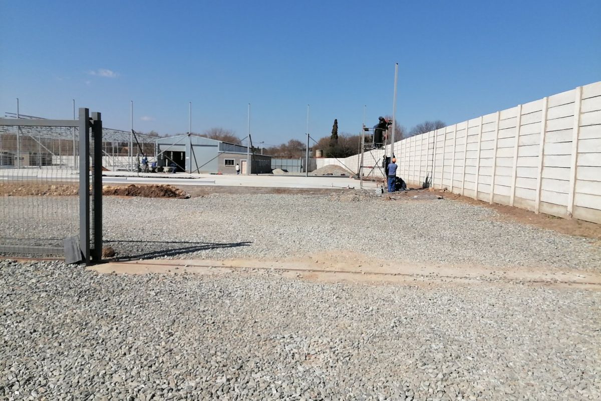 A vacant residential land plot under development, featuring a precast concrete boundary wall, gravel surface, and ongoing steel structural construction.