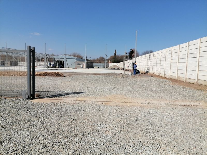 A vacant residential land plot under development, featuring a precast concrete boundary wall, gravel surface, and ongoing steel structural construction.