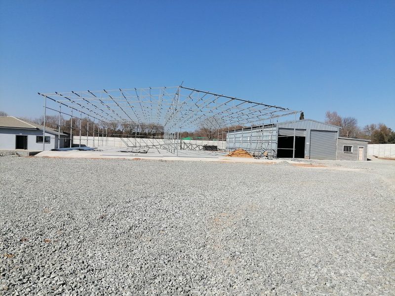 A view of a property under construction featuring a large steel frame structure, concrete slab, and an existing metal-clad building on a gravel lot.
