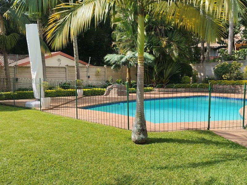A well-maintained outdoor pool area featuring a paved surround, safety fencing, and a lush, landscaped garden setting.