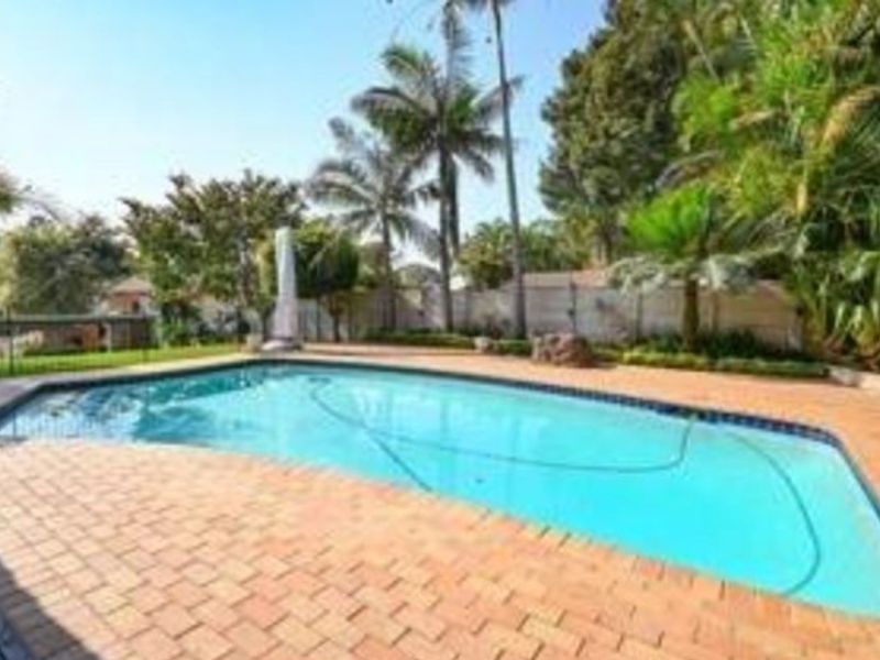 A well-maintained outdoor swimming pool area featuring brick paving, established garden landscaping, and a secure boundary wall.