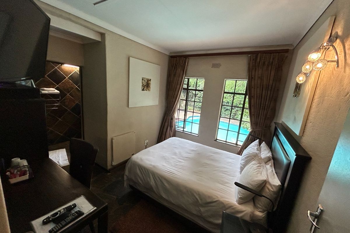 A cozy bedroom featuring an en-suite bathroom, tiled flooring, and windows overlooking a swimming pool area.