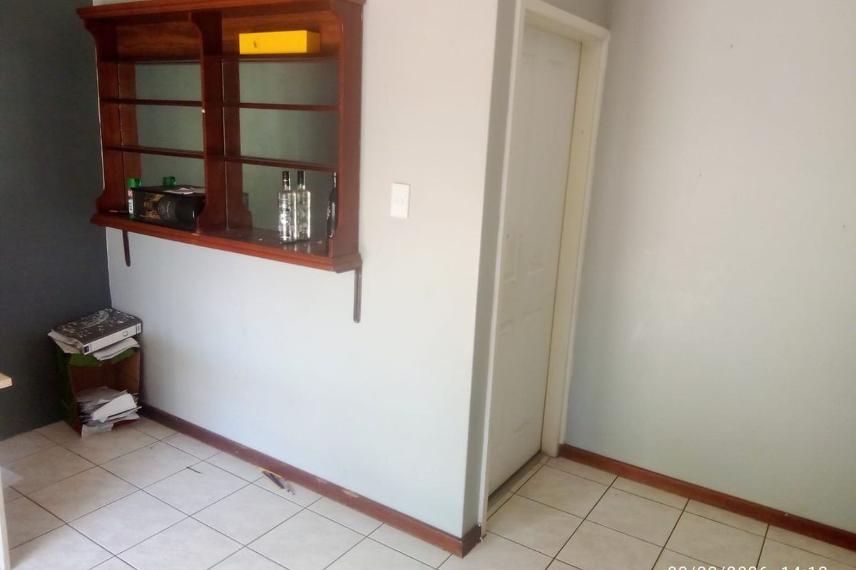 A sparsely furnished living area featuring tiled flooring, a wall-mounted wooden display shelf, and a standard internal door.